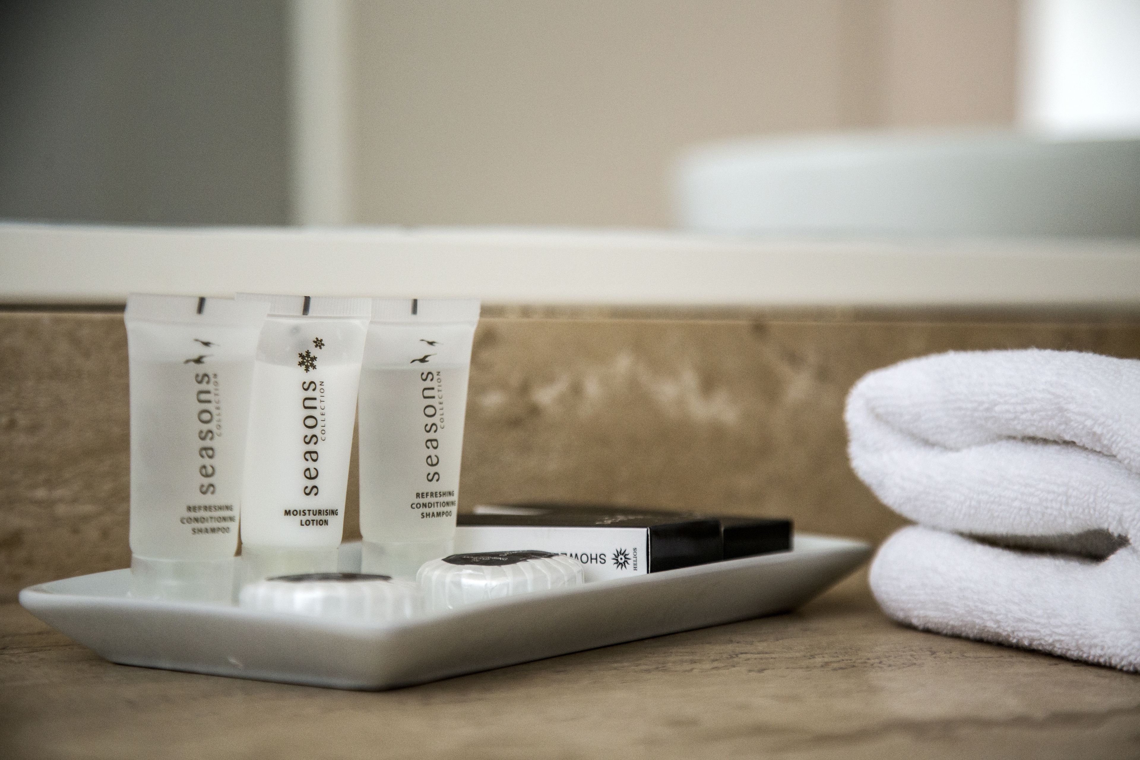 A tray with three tubes of Seasons Collection shampoo and conditioner, a black bar of soap, and a folded white towel on a beige countertop.
