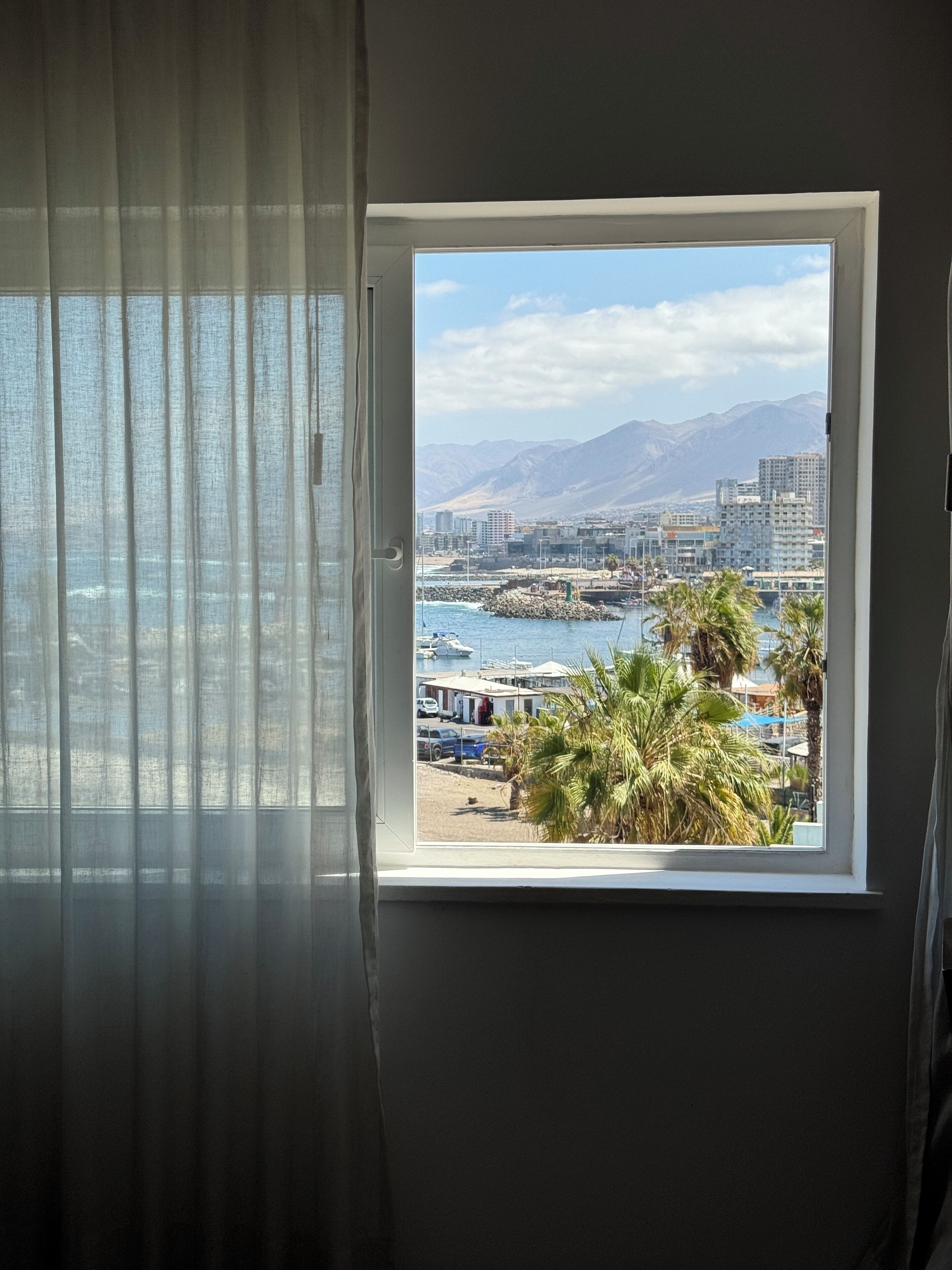 Ventana con vista a un puerto deportivo, barcos y un paisaje montañoso.