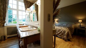 A bathroom with a sink, mirror, and window. The room has wooden flooring and a bed with a patterned bedspread. - Hotel Lafarques (Pepinster)