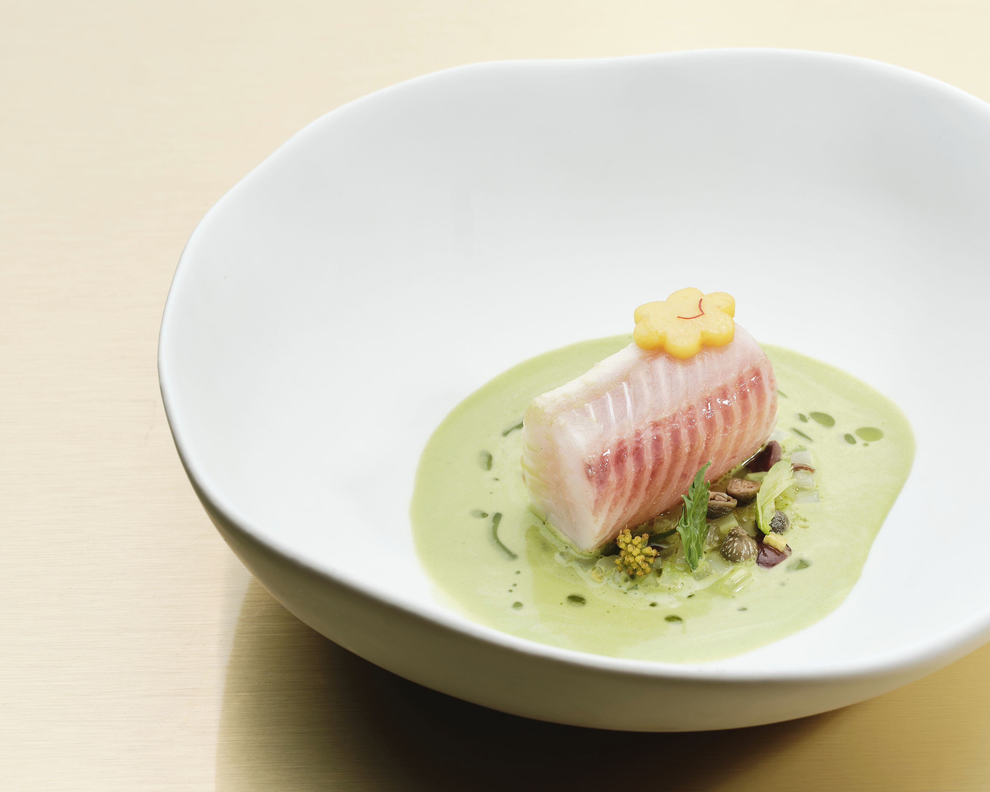 Een witte schaal met een gerecht bestaande uit een visfilet, versierd met een gele bloem en omringd door groene saus en kruiden.