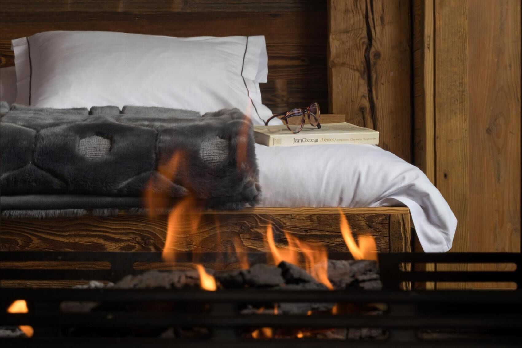 A bed with a white pillow and a gray blanket, a book titled 'Jean Cocteau Poems', and a pair of glasses on the bedside table.