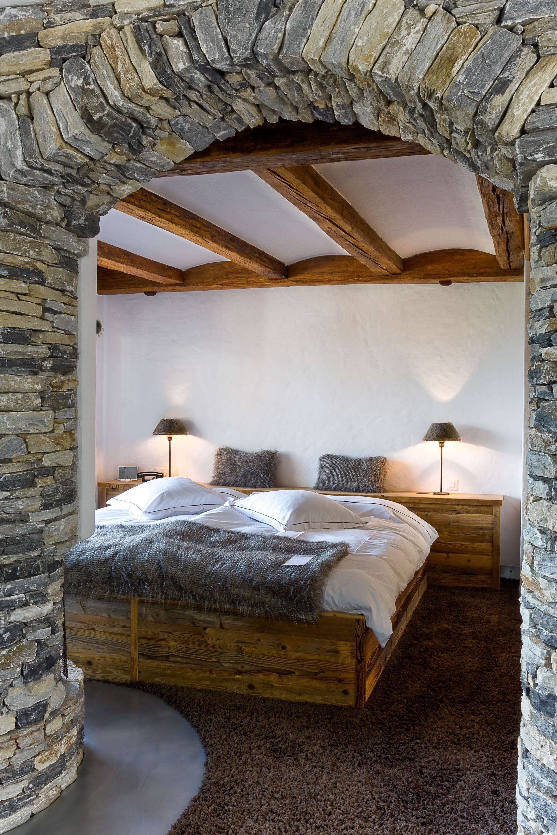 A bedroom with a stone archway, wooden beams, a bed with white linens, two nightstands with lamps, and a stone wall.