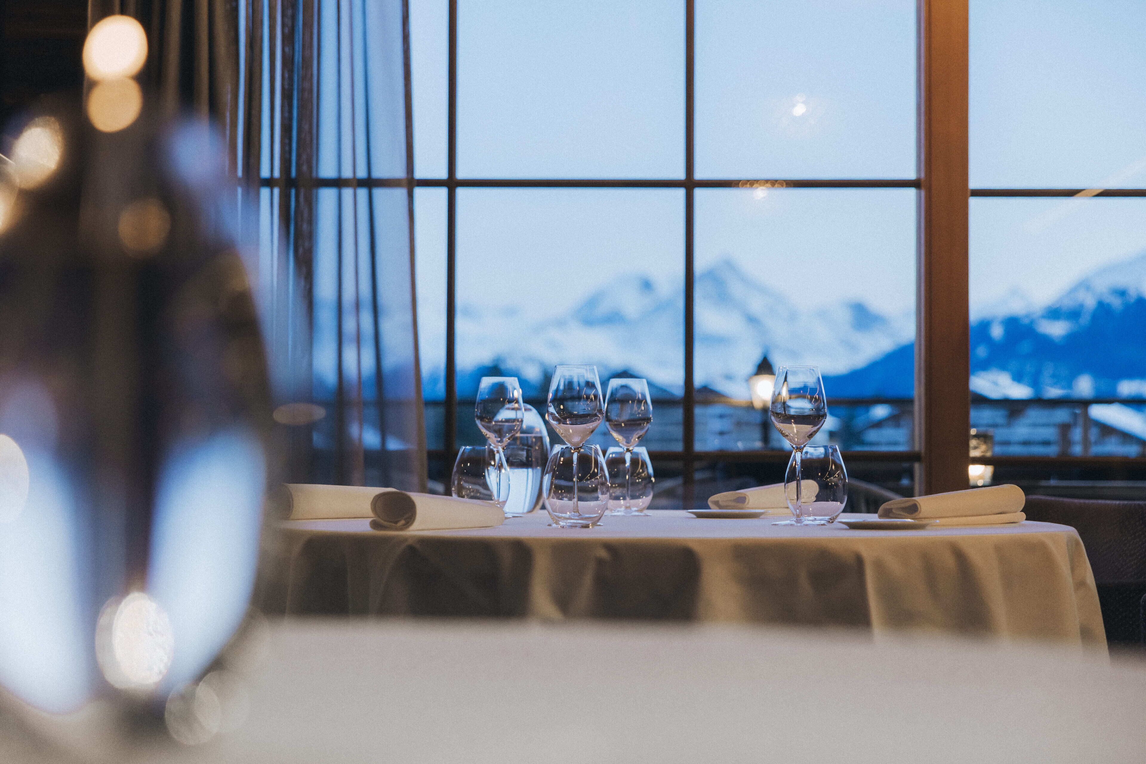 A dining table with multiple wine glasses and a menu, set against a backdrop of a mountain view through large windows.