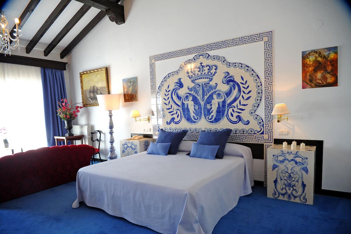 A bedroom with a large bed, a chandelier, a blue and white tiled headboard, and a red armchair.
