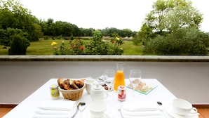 Une table de petit-déjeuner dressée avec de la vaisselle blanche, un panier de pain, un pichet de jus d’orange et une vue sur un jardin avec des arbres et des fleurs.
