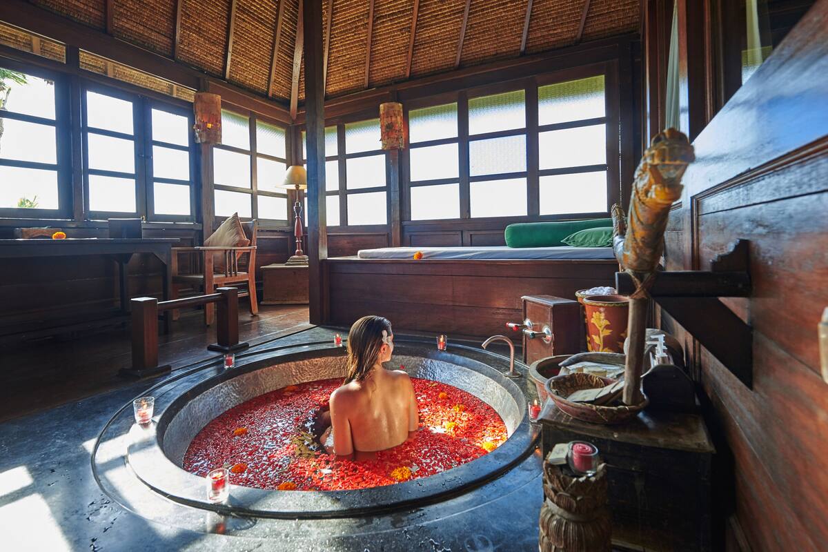 A person in a circular hot tub with a wooden interior, surrounded by windows and various amenities.