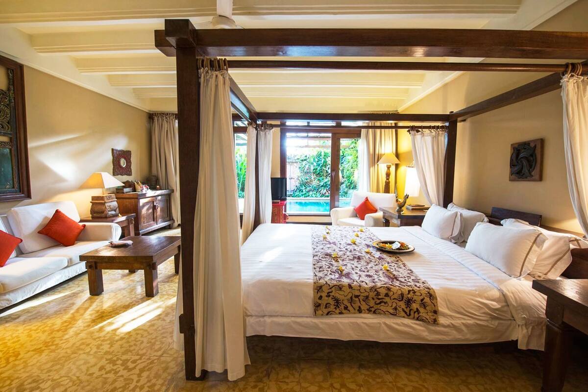 A four-poster bed with a view of a pool, a sofa, a nightstand, and a painting on the wall.