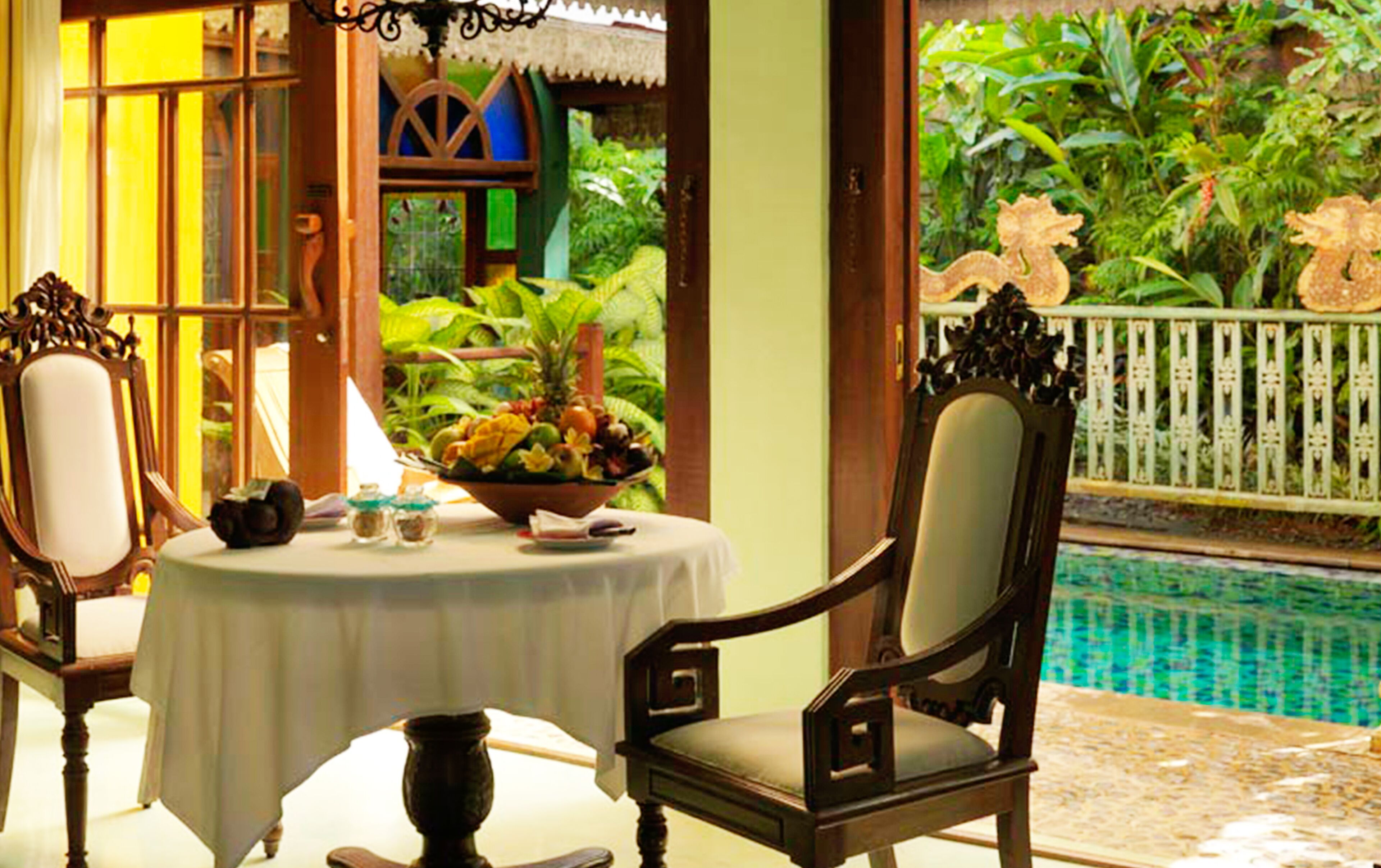 A dining area with a table set for two, a decorative bowl of fruit, and a view of a swimming pool and lush greenery.
