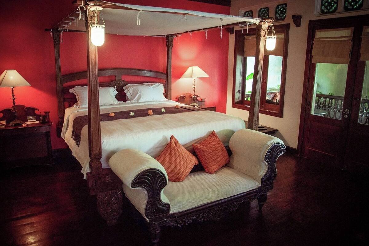 A four-poster canopy bed with white linens and multiple pillows, a wooden nightstand with a lamp, and a chair with cushions.