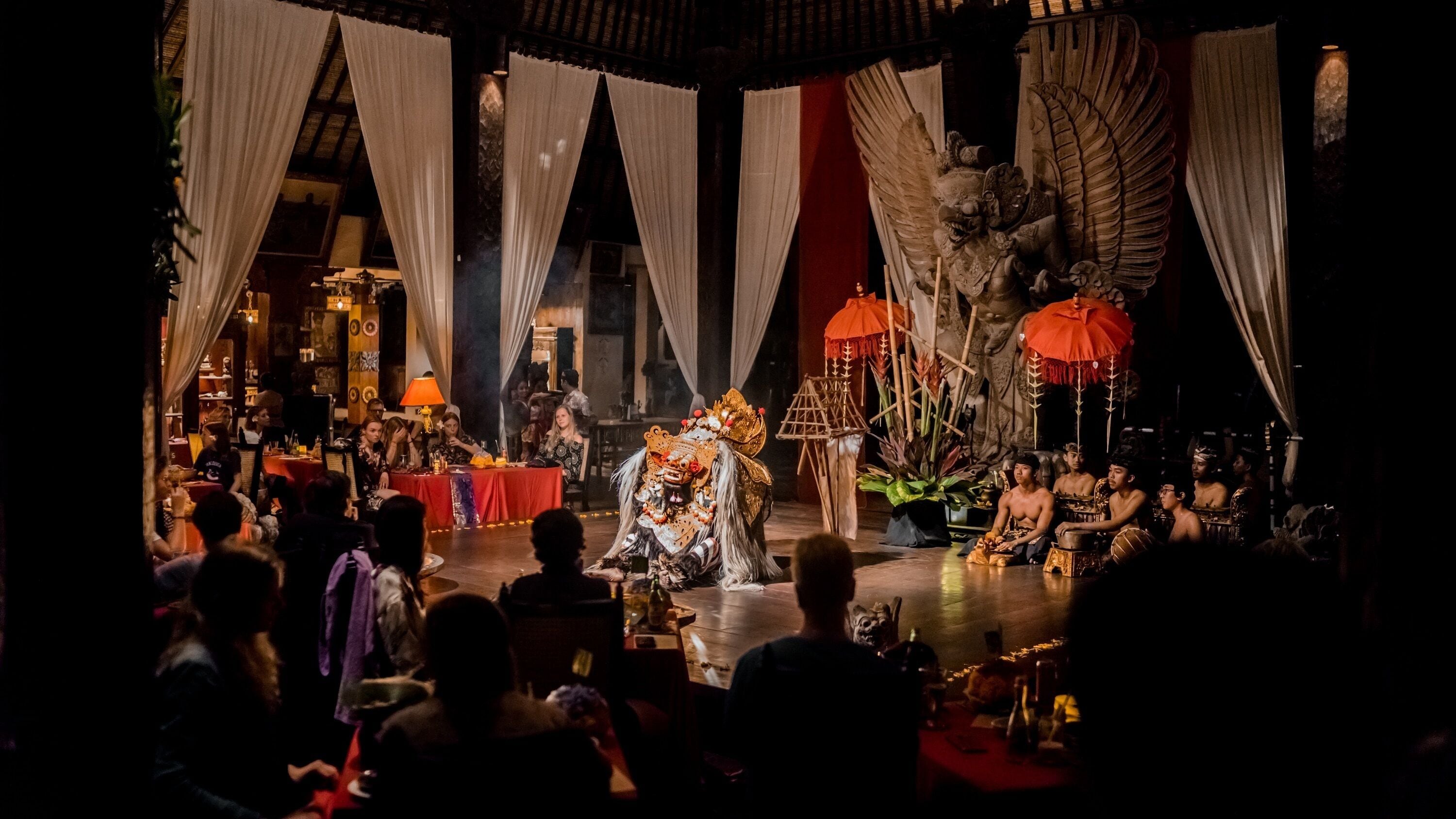 Un escenario con un artista en un elaborado vestuario, un público sentado en hileras y un telón de fondo con elementos arquitectónicos.
