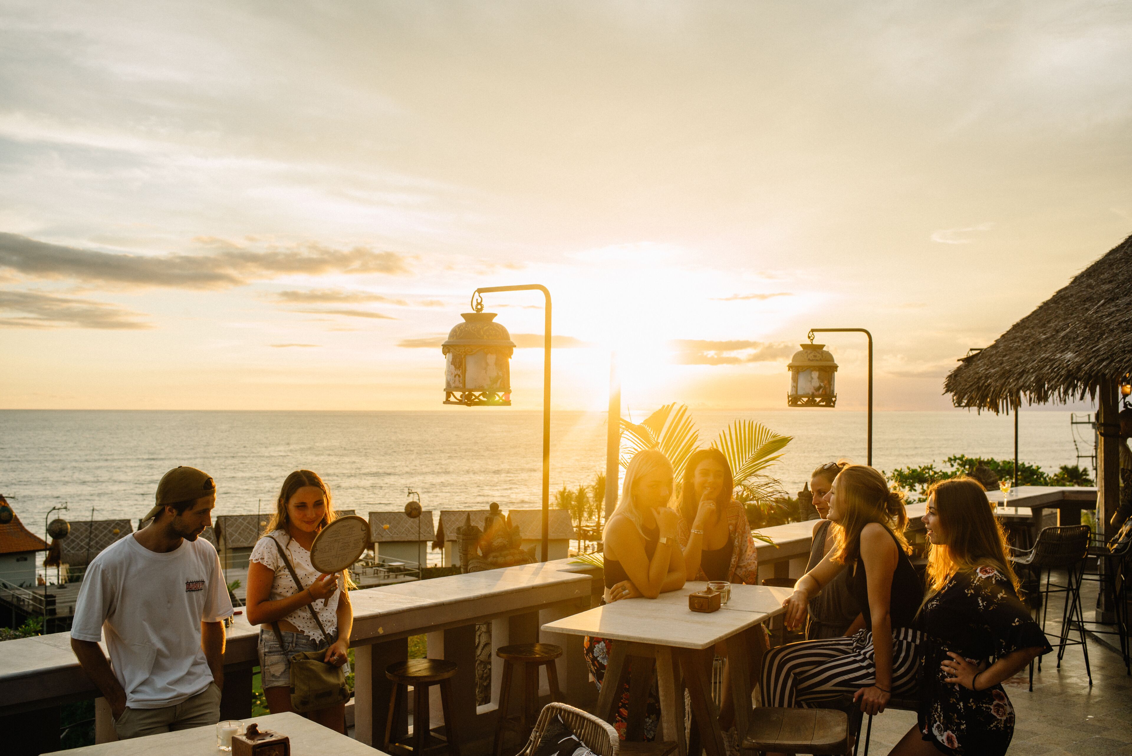 Un restaurant en bord de mer avec une terrasse offrant une vue sur l’océan, comprenant des tables et des chaises, et une structure au toit de chaume en arrière-plan.