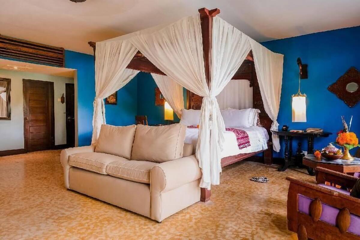 A room with a four-poster bed, a sofa, a small table with a vase, and a wall-mounted lamp.