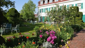 Un grand bâtiment à la façade blanche et aux volets verts, entouré d’un jardin bien entretenu où poussent différentes espèces de fleurs, et doté d’une terrasse pavée avec des meubles pour s’asseoir à l’extérieur.