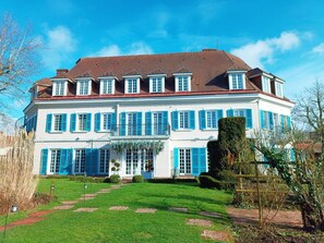 Un edificio grande de dos pisos con una fachada simétrica, múltiples ventanas con contraventanas azules y un césped bien cuidado con un camino de ladrillo que conduce a la entrada.