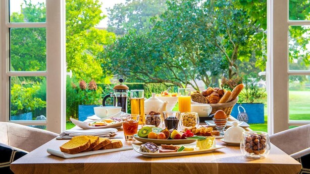 Une table de petit-déjeuner dressée avec divers aliments et boissons, le tout mis en valeur par un décor de verdure luxuriante visible à travers de grandes fenêtres.