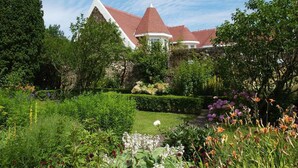 Une grande maison avec un toit rouge et une tour, entourée de jardins luxuriants et d’arbres.
