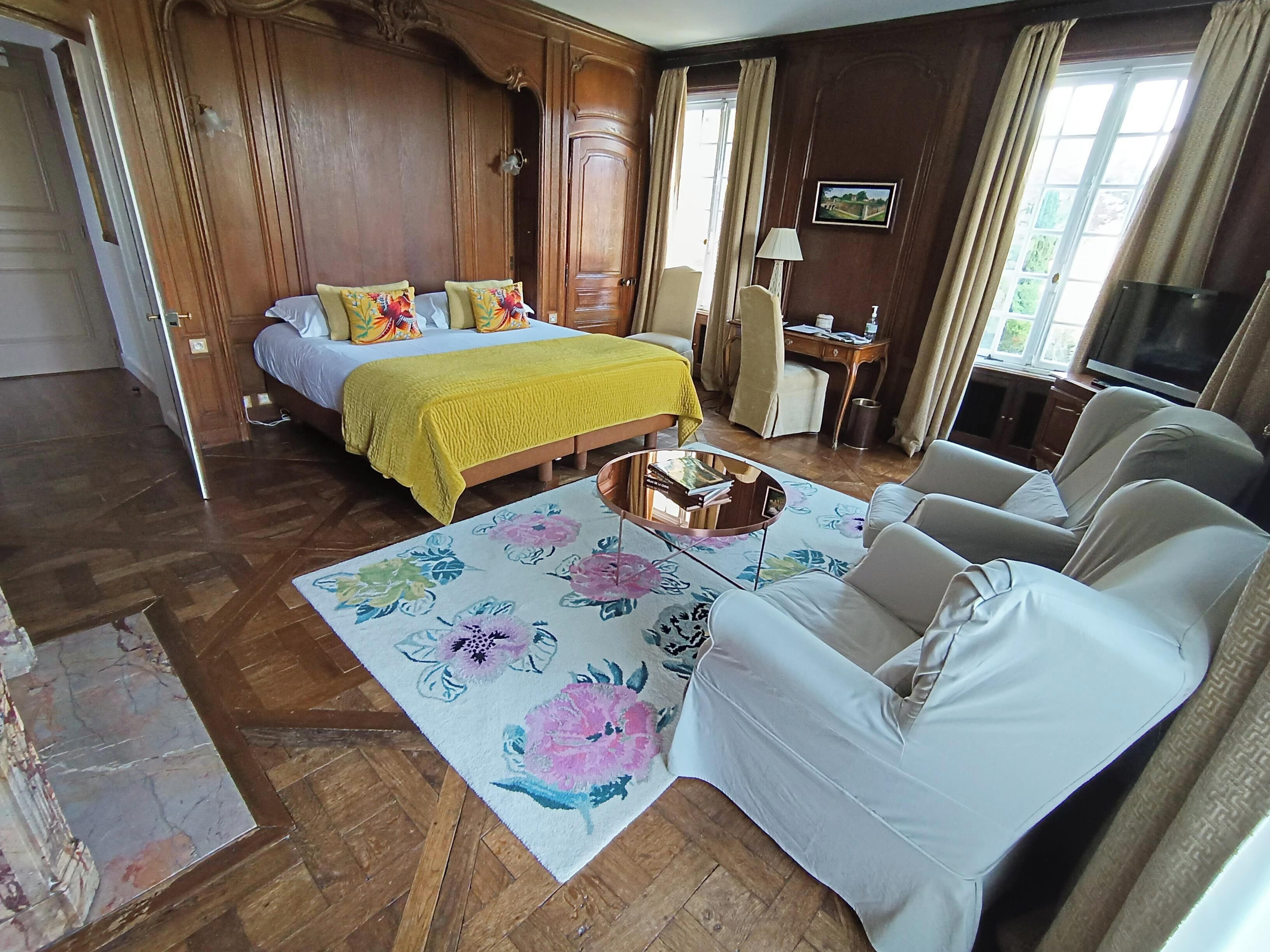 Habitación de hotel con cama, sofá, mesita de centro, televisor y ventana con cortinas.