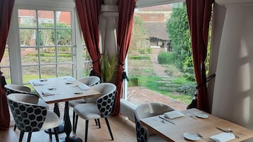 A dining area with a table set for four, surrounded by hexagon-patterned chairs, large windows with red curtains, and a view of a garden.
