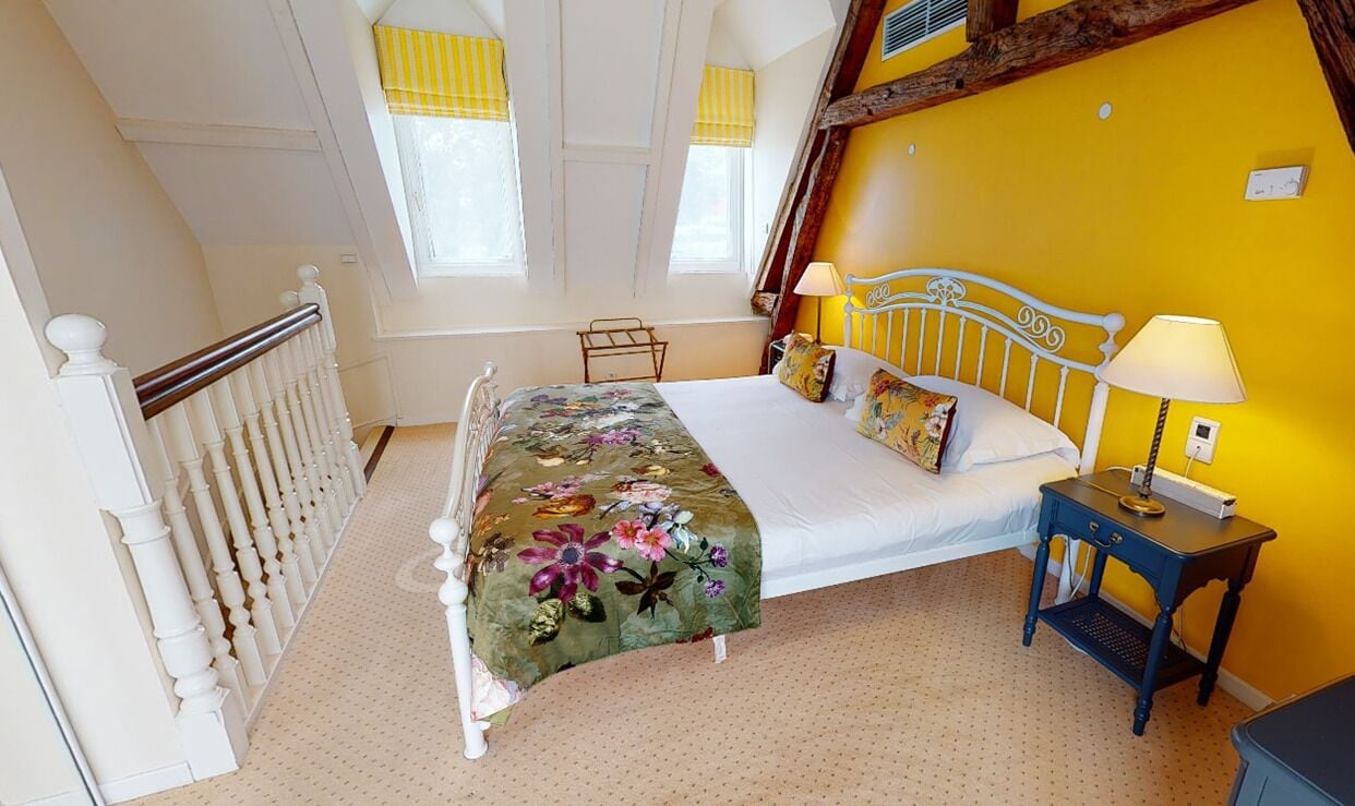 Quarto com cama, mesa de cabeceira, janela com cortinas amarelas e vigamento de madeira no teto.