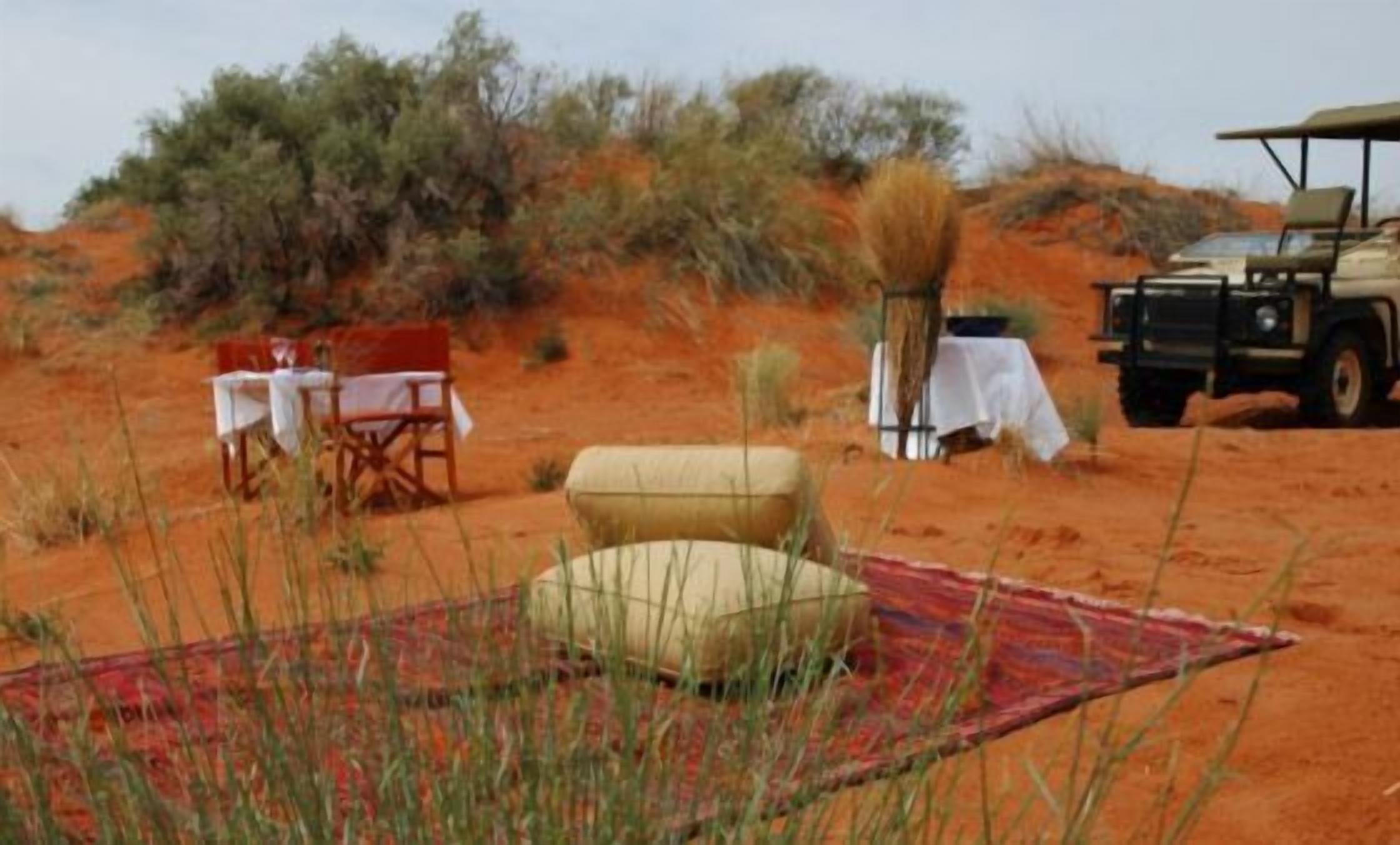 Paisaje desértico con una mesa y sillas, un espacio de descanso cubierto y un vehículo.