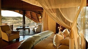 A bedroom with a large bed, a wooden desk, and a chair, with a view of the outdoors. - The Motse - Tswalu Kalahari Luxury Private Game Reserve (Kuruman)