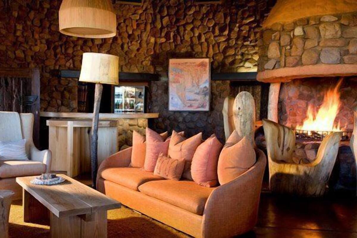 A cozy lounge area with a stone wall, a wooden sofa with cushions, a wooden table, and a fireplace with a visible fire.