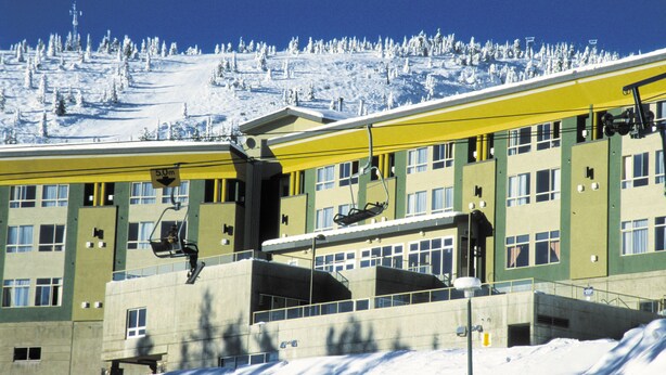 A multi-story building with a ski lift, surrounded by snow-covered trees and a clear blue sky. - Inn at Big White (Beaverdell)