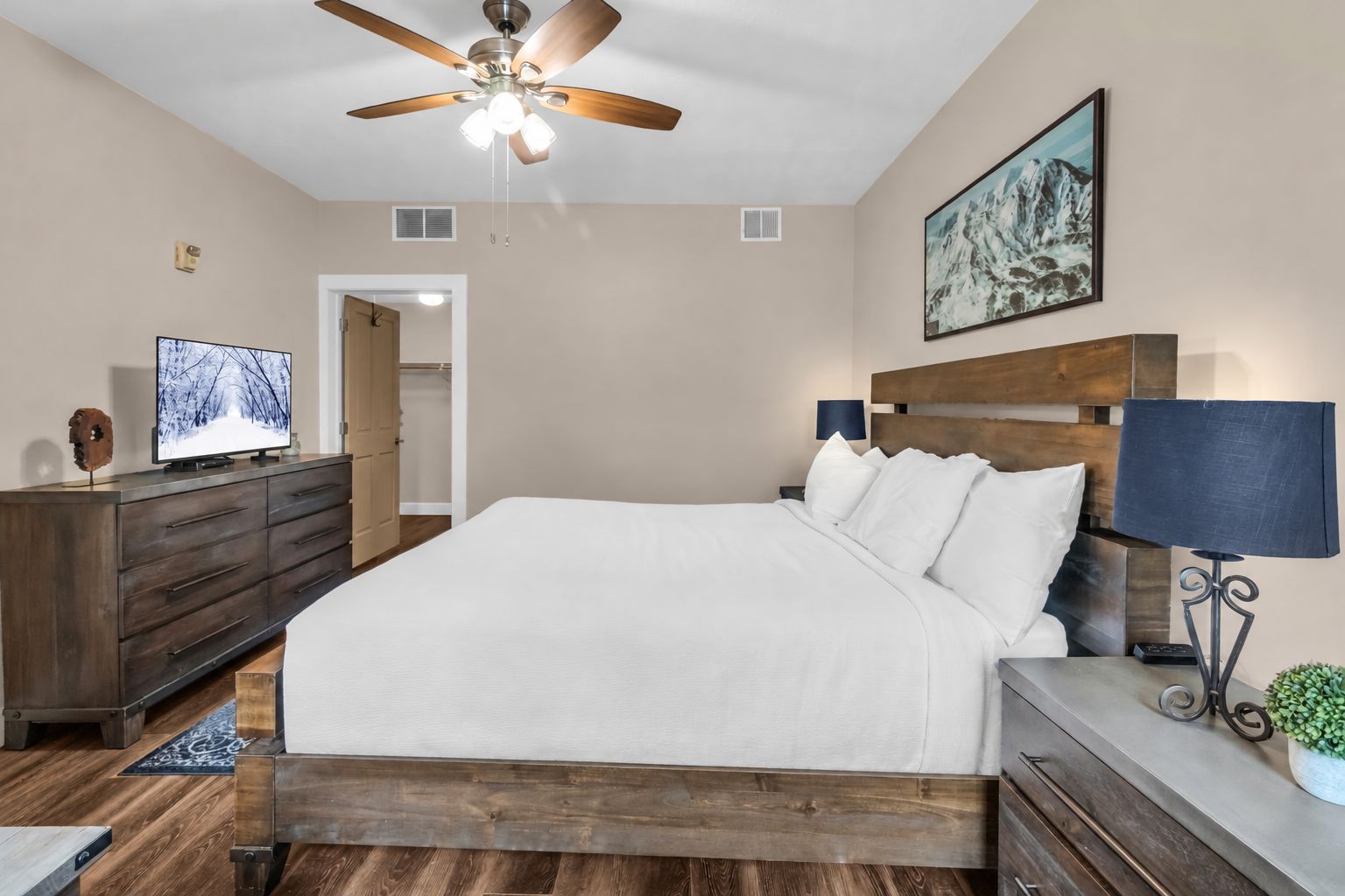 Une chambre à coucher avec un lit, une tête de lit en bois, un ventilateur de plafond, une télévision, une commode et un tableau au mur.