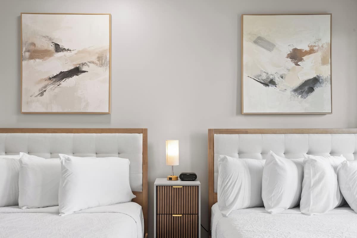 A bedroom with two beds, each with a wooden headboard, a bedside table with a lamp, and abstract paintings on the wall.