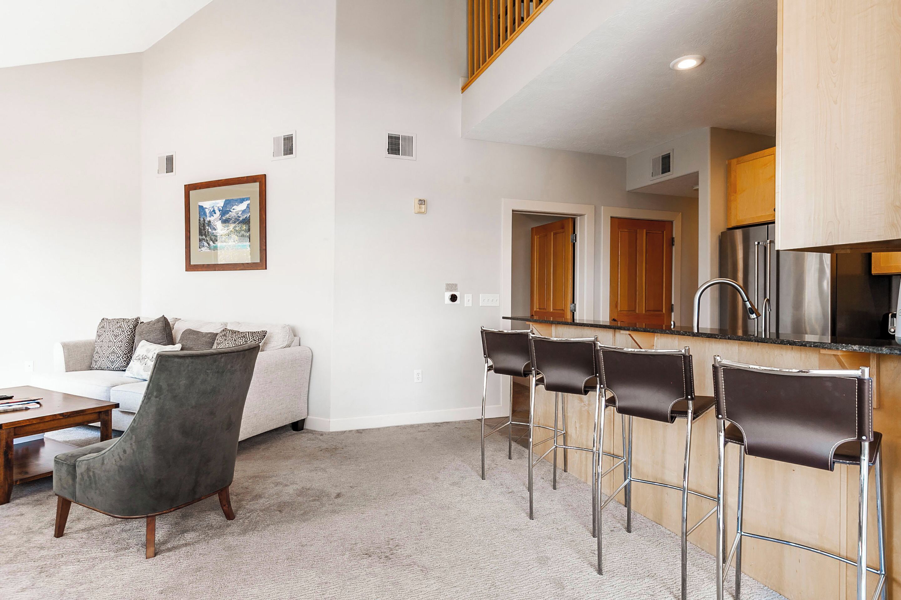 A modern living room with a grey armchair, a wooden coffee table, and a white sofa with patterned cushions. There are two black bar stools and a kitchen area with a stainless steel refrigerator.