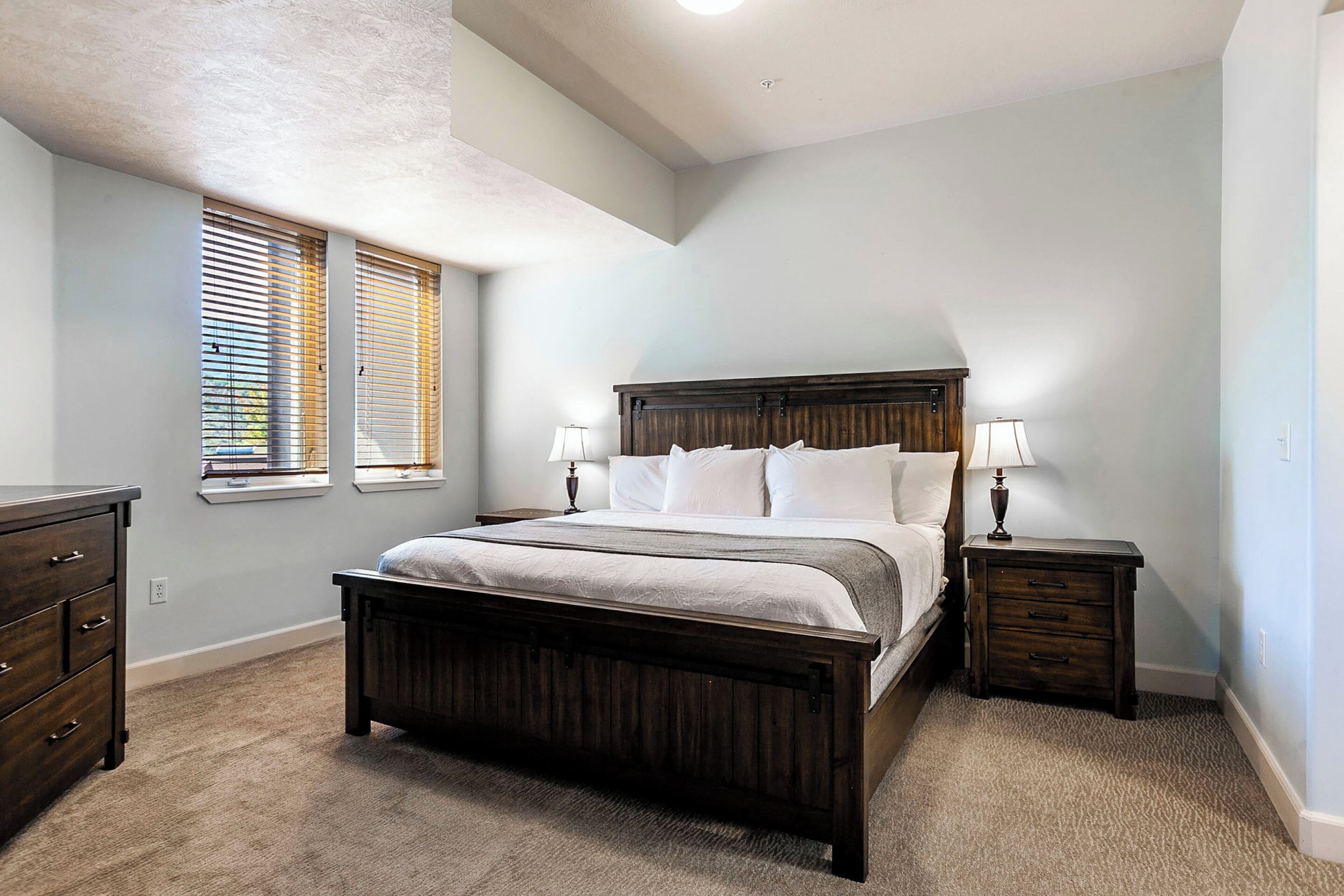 A bedroom with a large bed, two bedside tables with lamps, a dresser, and a window with blinds.