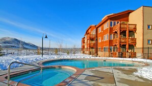A swimming pool with a wooden deck, surrounded by a metal fence, and a building with balconies in the background. - Crestview Condominiums by All Seasons Resort Lodging (Park City)