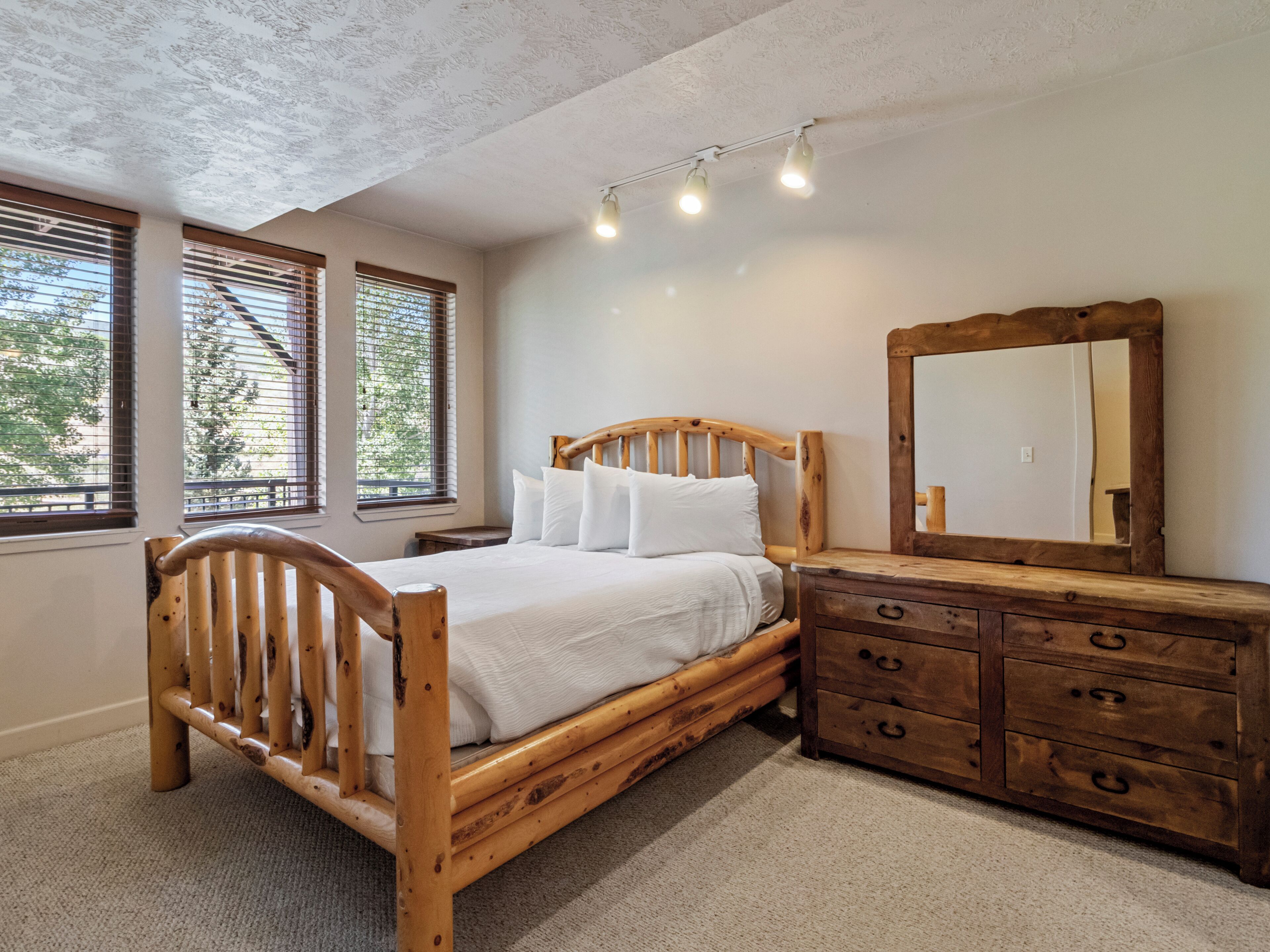 A bedroom with a wooden bed, a dresser, a mirror, and windows with blinds.