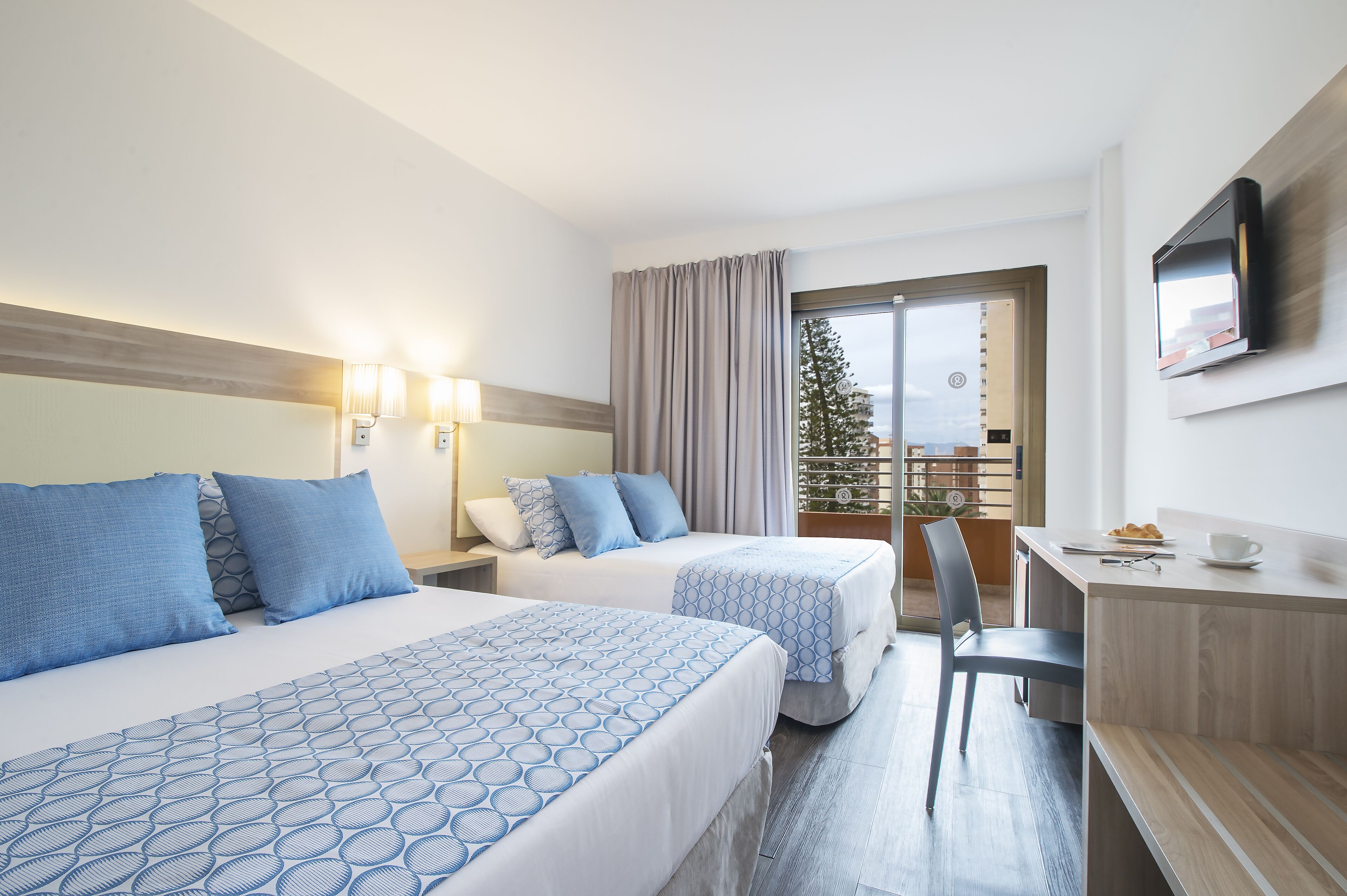 Habitación de hotel con dos camas, un escritorio, una silla, un televisor y un balcón con vista a un árbol.