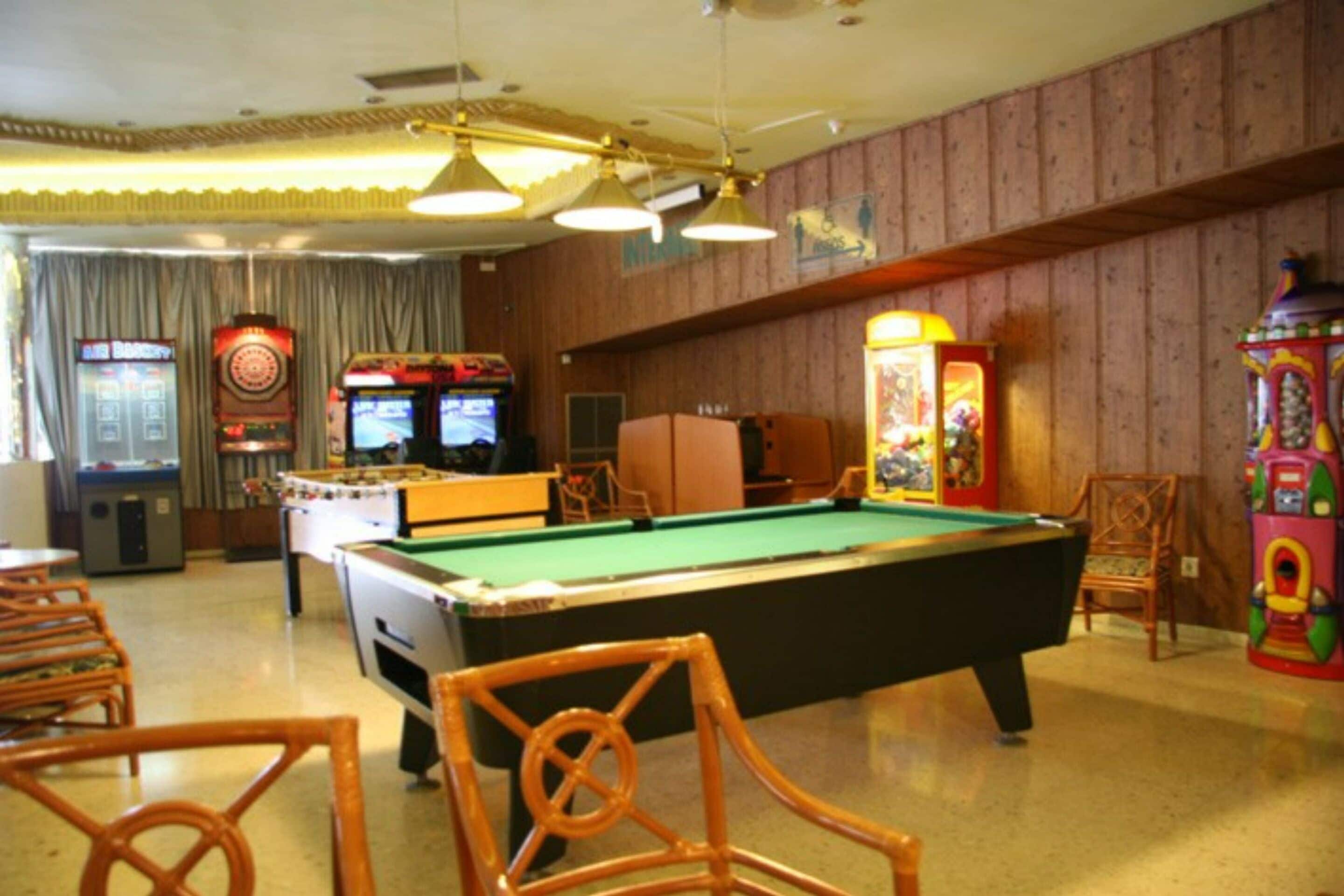 A pool table with surrounding chairs, arcade games, and a jukebox in a room with wooden paneling and ceiling lights.