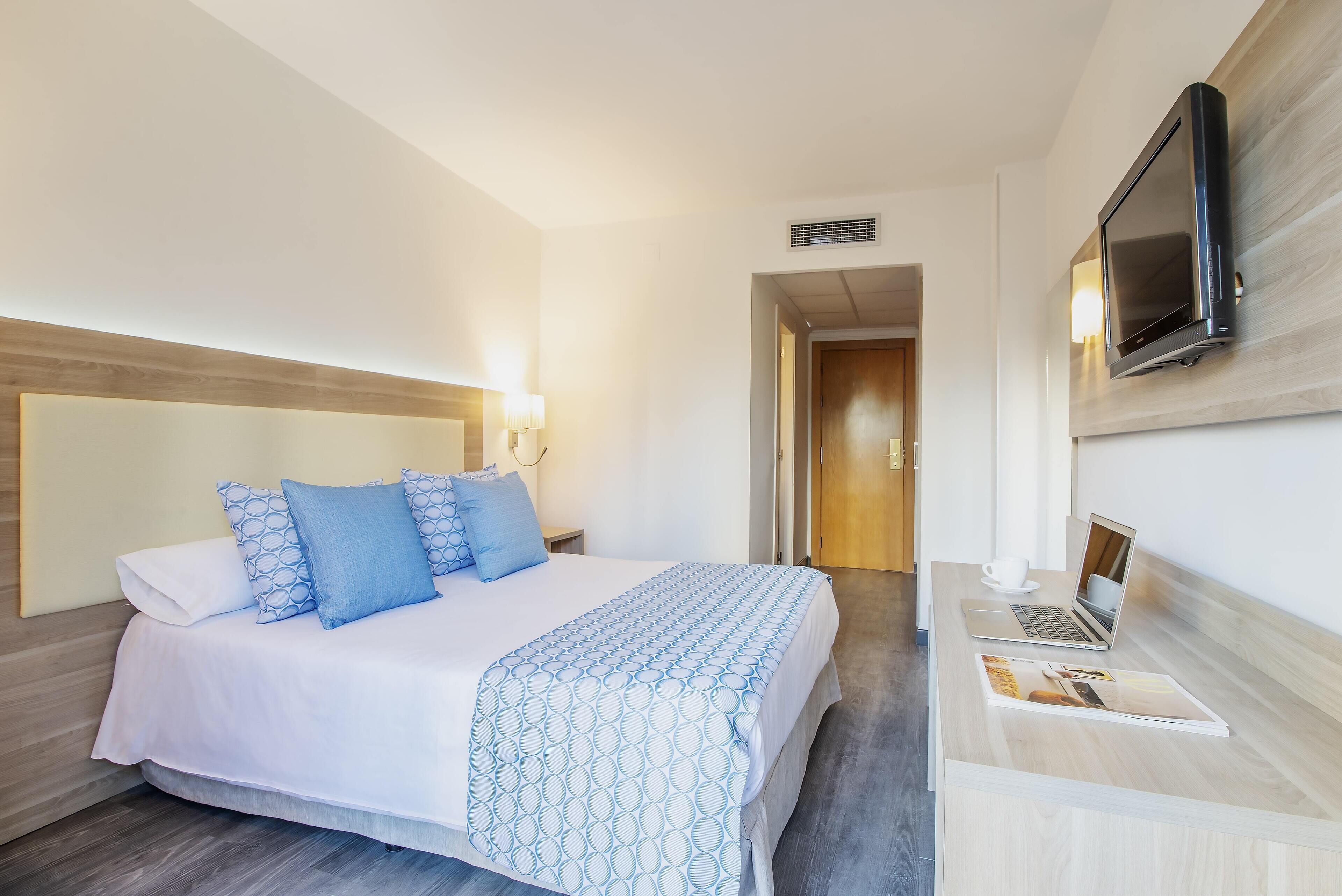Una habitación de hotel con una cama, un escritorio con una computadora portátil y un televisor montado en la pared.