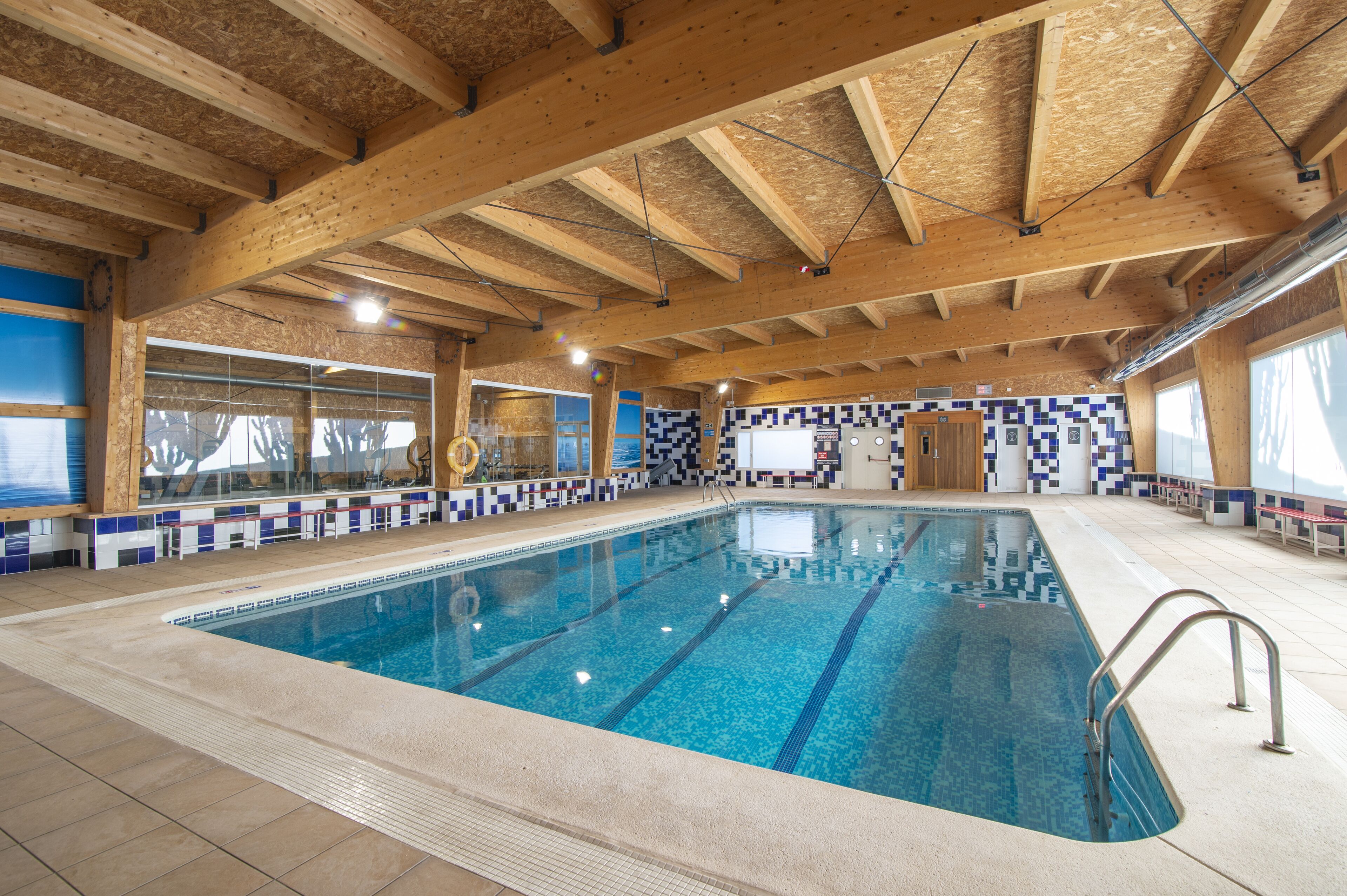 An indoor swimming pool with a tiled border, wooden beams, and a blue-tiled wall.