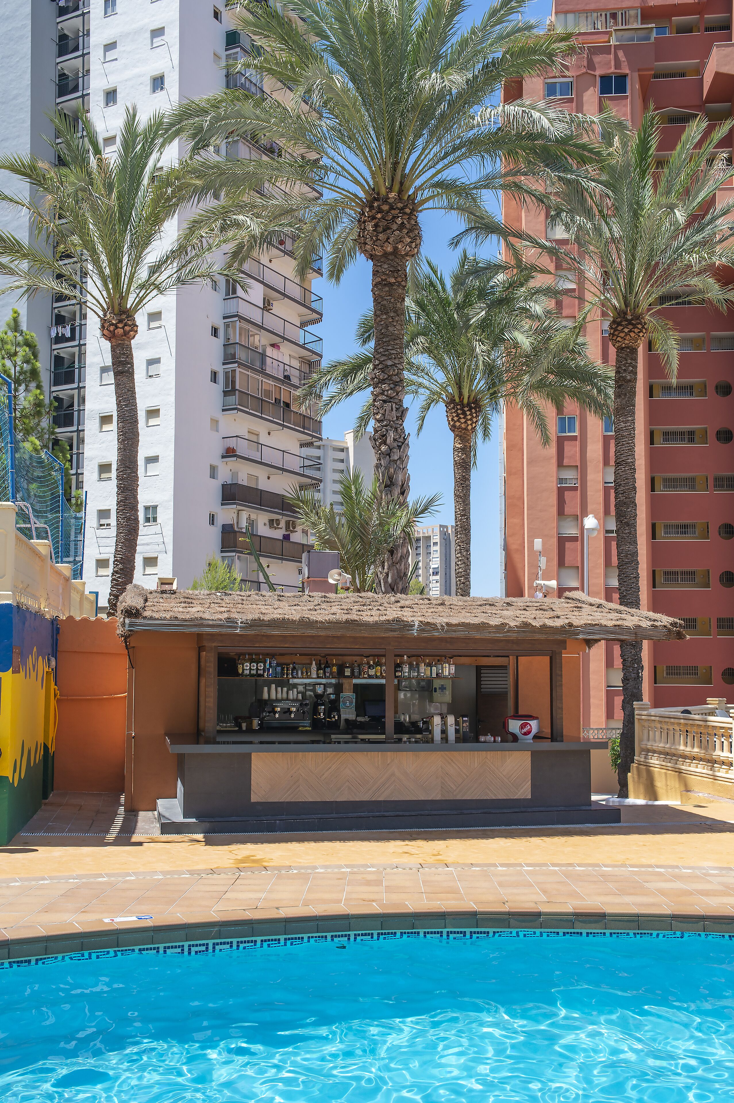 Un bar junto a la piscina con techo de paja, rodeado de palmeras y edificios altos.