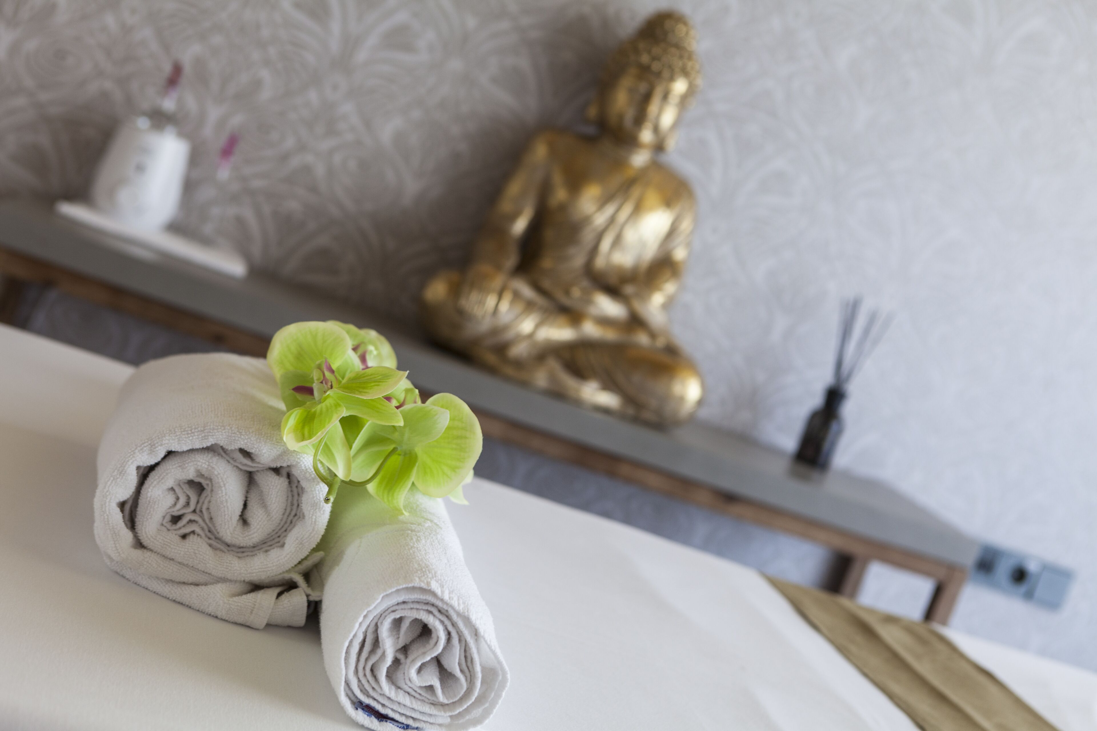 A neatly made bed with rolled towels and a small green plant. A golden Buddha statue is visible in the background.