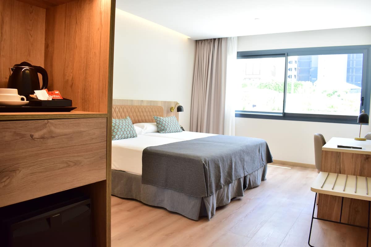 A hotel room with a bed, a desk, a chair, a coffee maker, and a window with a view of buildings.