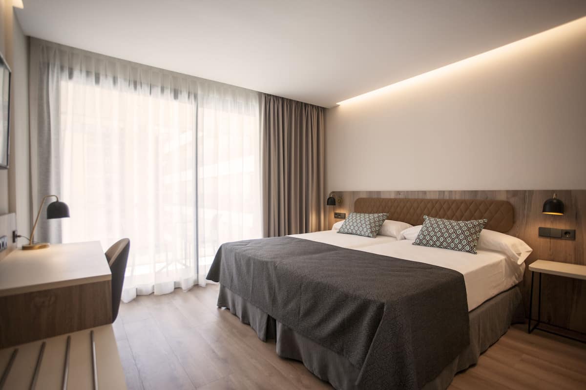 A modern hotel room with a large bed, bedside tables, a desk, and a window with curtains.