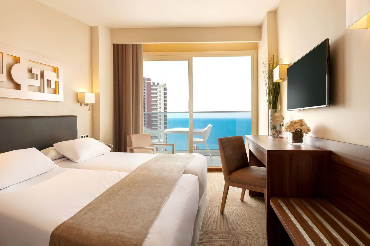 A hotel room with two beds, a desk, a chair, a TV, and a balcony with a view of the ocean.