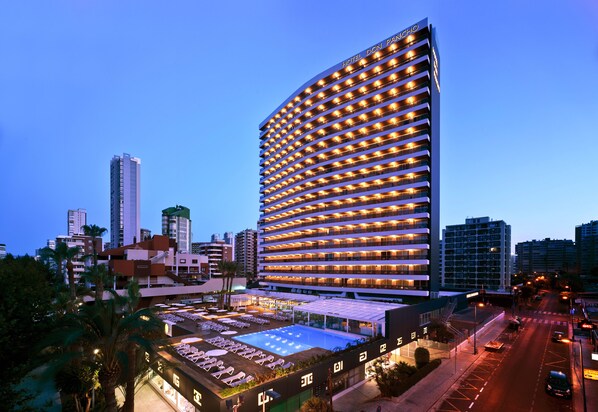 Un hotel de altura con fachada curva, varios balcones y zona de piscina en la azotea.