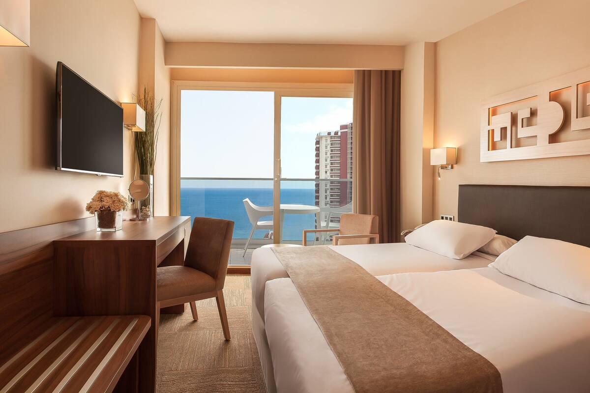 A hotel room with a bed, a desk, a chair, a TV, and a view of the ocean.