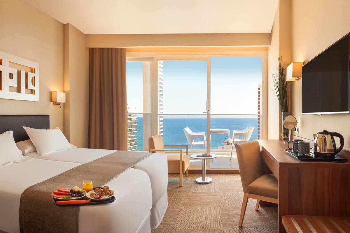 A hotel room with a large bed, a desk with a chair, a small table with a chair, and a balcony with a view of the ocean.