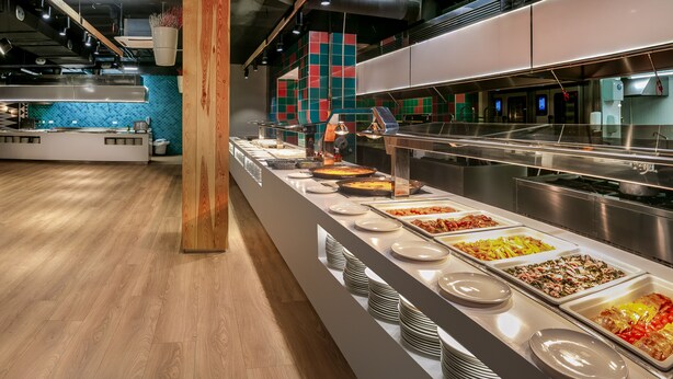 Zona de bufé con diversas opciones de comida, un pilar de madera y una cocina moderna al fondo.
