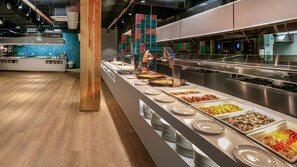 Un espace buffet proposant divers plats, un pilier en bois et, au fond, une cuisine moderne.