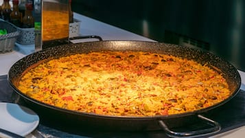 Una sartén grande, redonda y negra llena de un plato que parece ser una preparación a base de arroz o cereal, posiblemente una paella u otro plato similar, con ingredientes visibles como aceitunas y pimientos rojos.