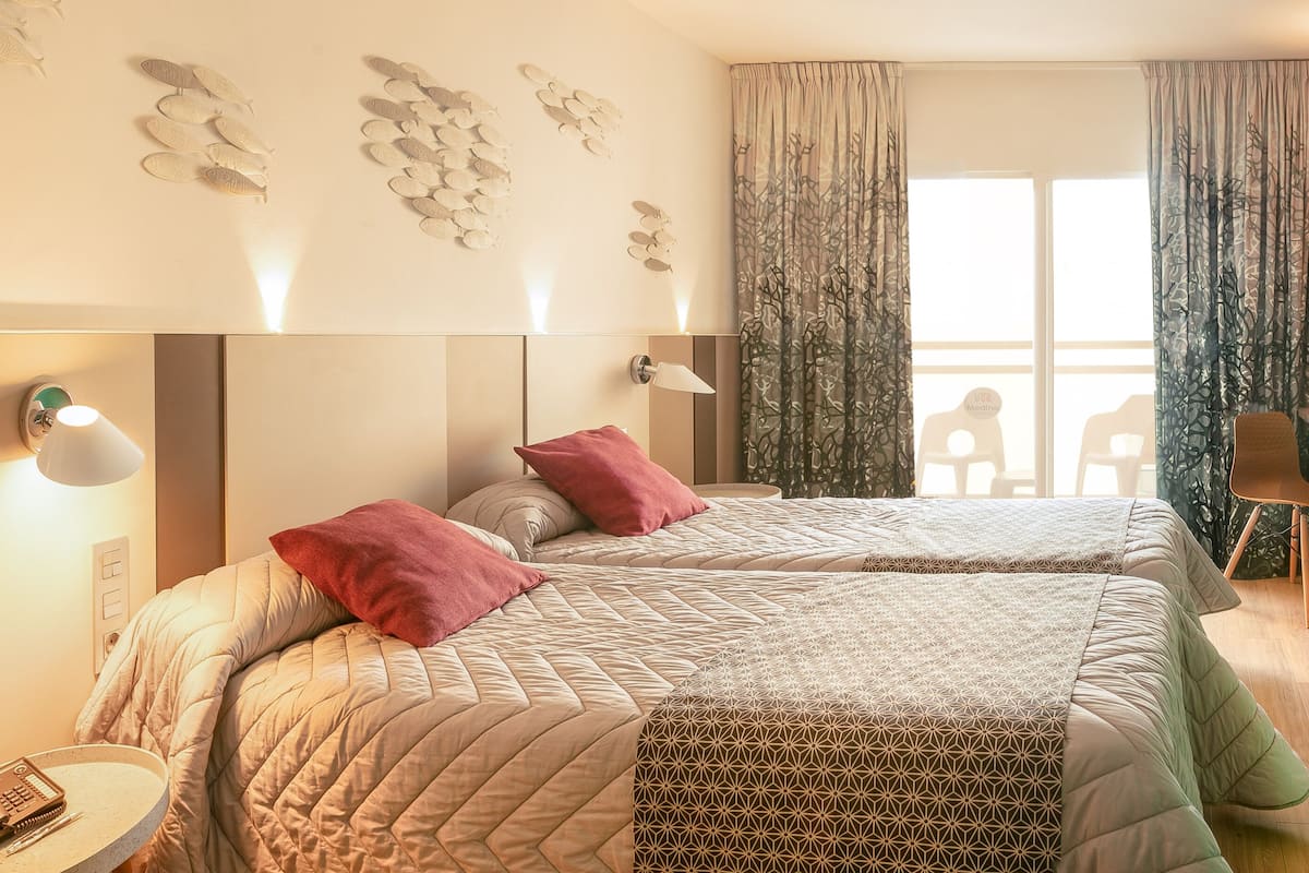 A hotel room with two beds, a TV mounted on the wall, a desk with a chair, and a window with curtains.