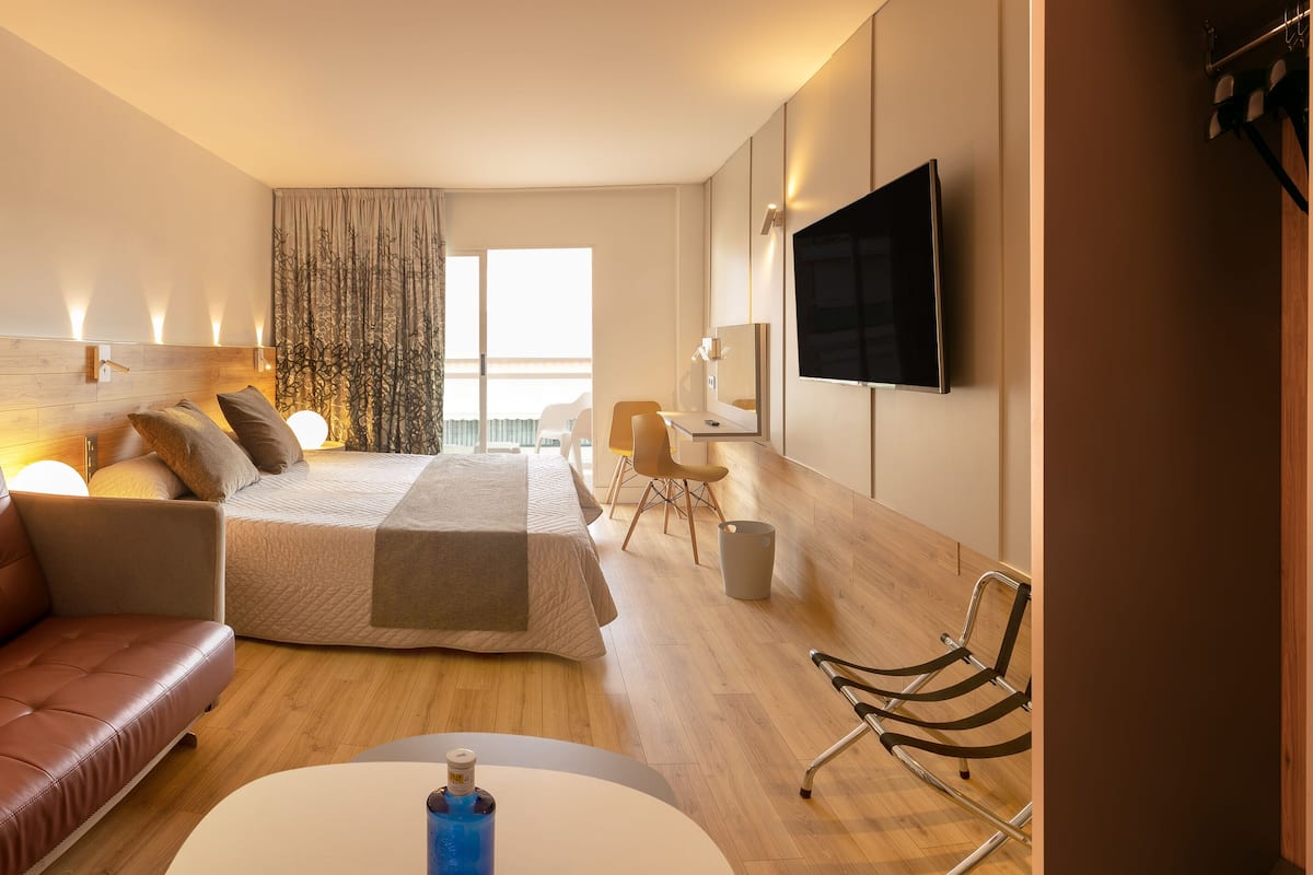 A modern hotel room with a leather sofa, a bed with a wooden headboard, a flat-screen TV, and a desk with a chair.